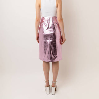 A woman shown from the back wearing a sleeveless white top and a shiny metallic pink skirt, standing against a plain light beige studio background.