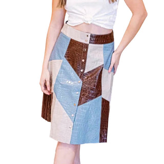 A woman models a patchwork leather skirt with blue, brown, and beige panels, paired with a white crop top and white sneakers in an indoor setting with pink curtains.