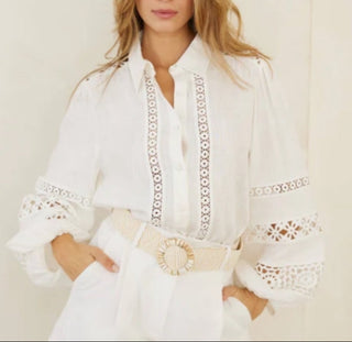 A woman modeling a white long-sleeve blouse with intricate lace and crochet embroidery on the front placket, cuffs, and shoulders, paired with a matching white belt featuring a large round decorative buckle.